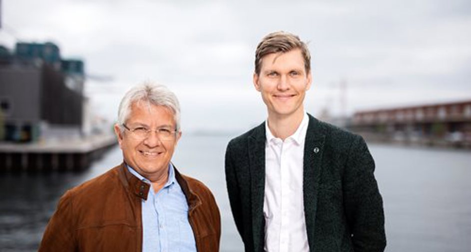 Billede af Lasse Frimand Jensen og DANVA´s formand Carsten Nystrup Lasse Frimand Jensen og Carsten Nystrup i brun jakke og mørk jakkesæt står smilende foran en havn med vand og bygninger i baggrunden.