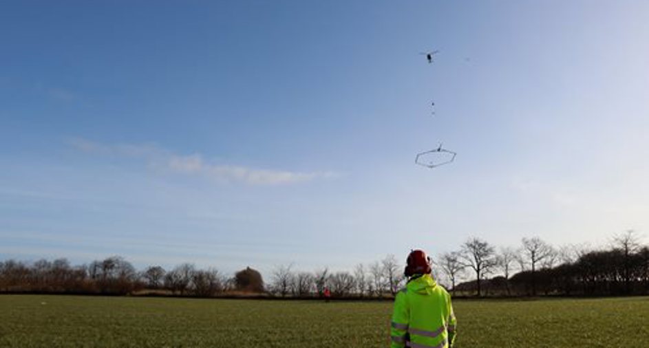Billede af mand der tester SkyTEM på en mark FOTO: SkyTEM: En person iført en neongul jakke og hjelm står på en mark og observerer en drone, der flyver i luften med et sekskantet instrument hængende under den.